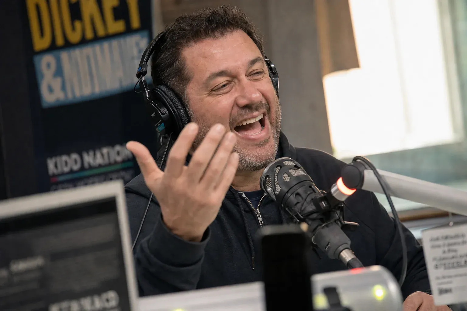 Radio host at the microphone caught mid-laugh during a live Father's Day break, gesturing animatedly with headphones on, capturing the debate-and-banter energy of Father's Day radio content