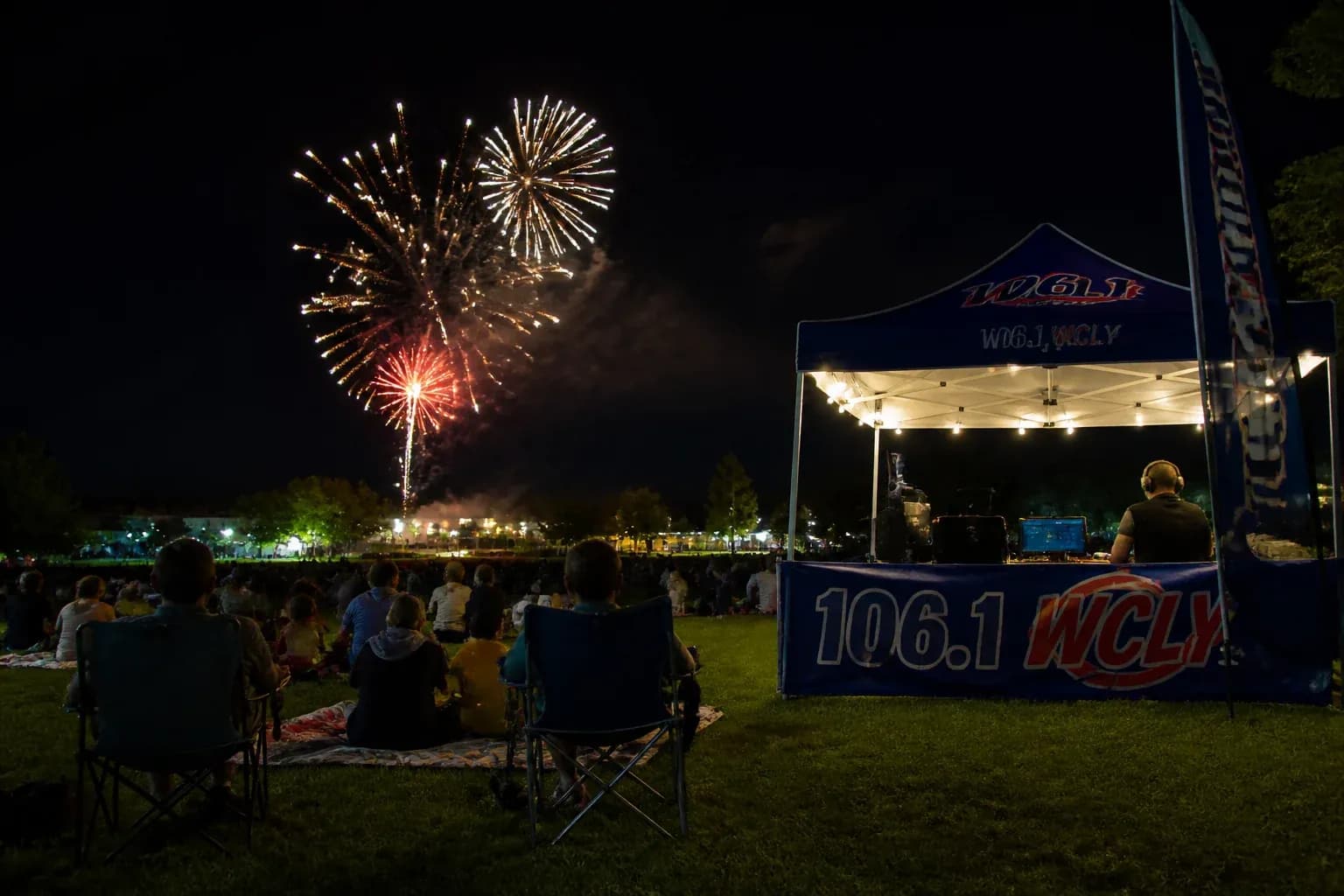 Community Fourth of July fireworks display over a small-town park at night, families watching with a radio station remote tent lit up nearby