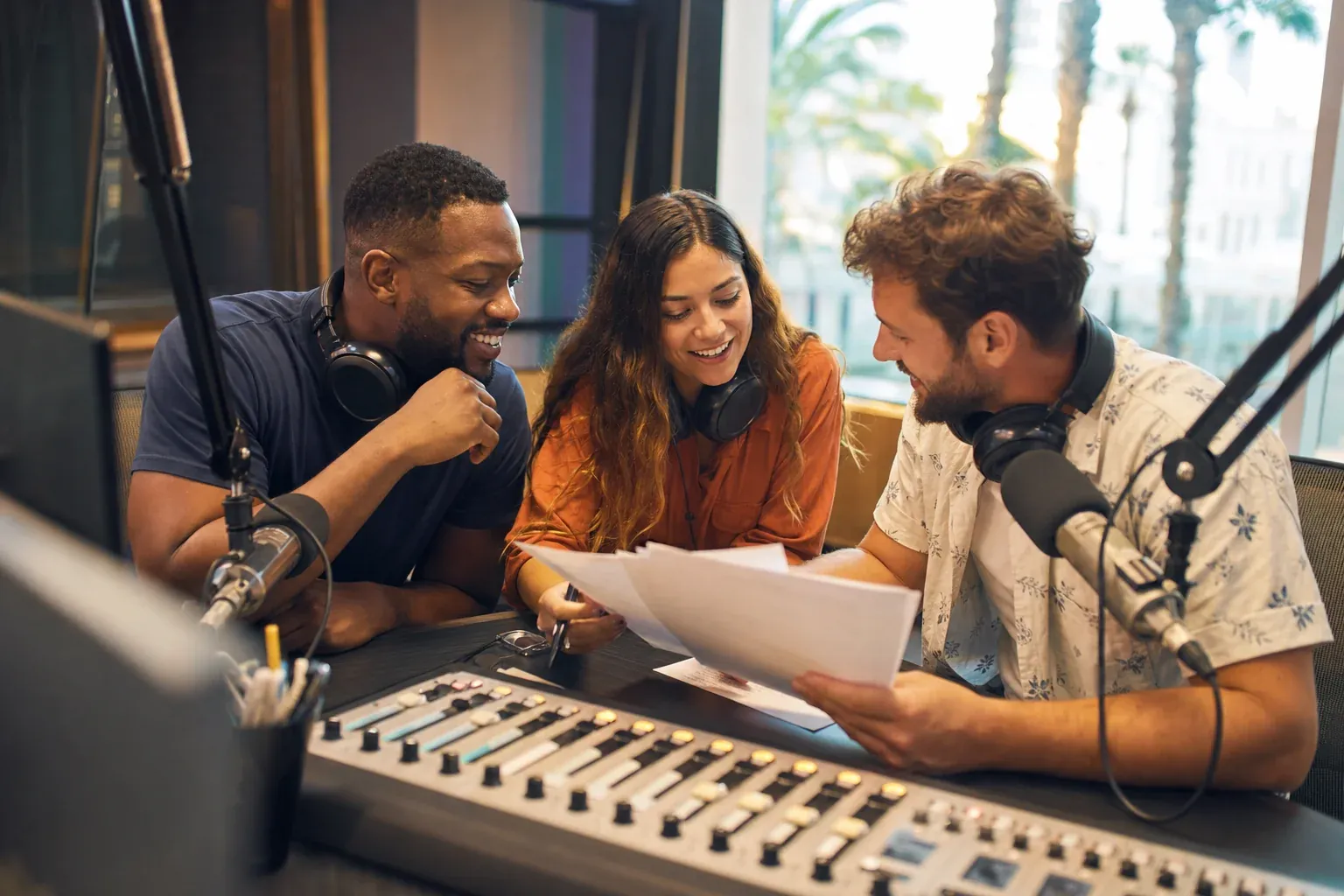 A morning show team at the studio console reviewing summer segment notes together, relaxed and energized in warm natural light