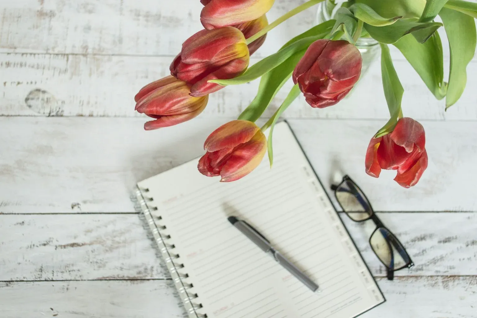 Open notepad, pen, reading glasses, and fresh tulips on a bright white wooden desk in warm natural light — a spring planning workspace