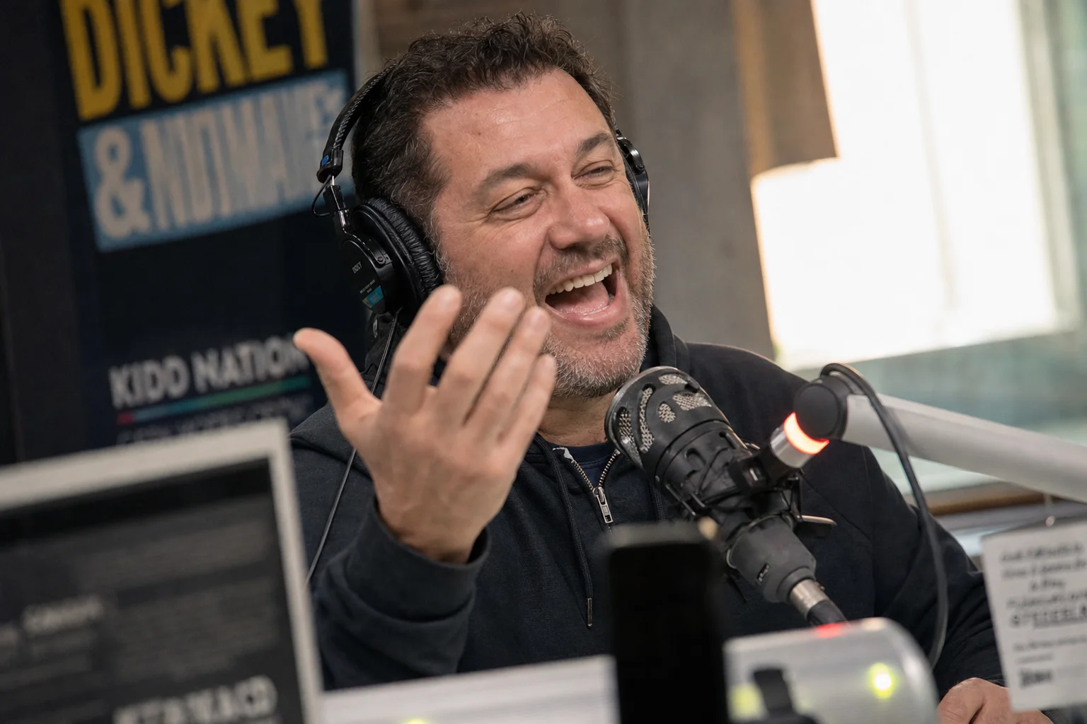 Radio host at the microphone caught mid-laugh during a live Father's Day break, gesturing animatedly with headphones on, capturing the debate-and-banter energy of Father's Day radio content
