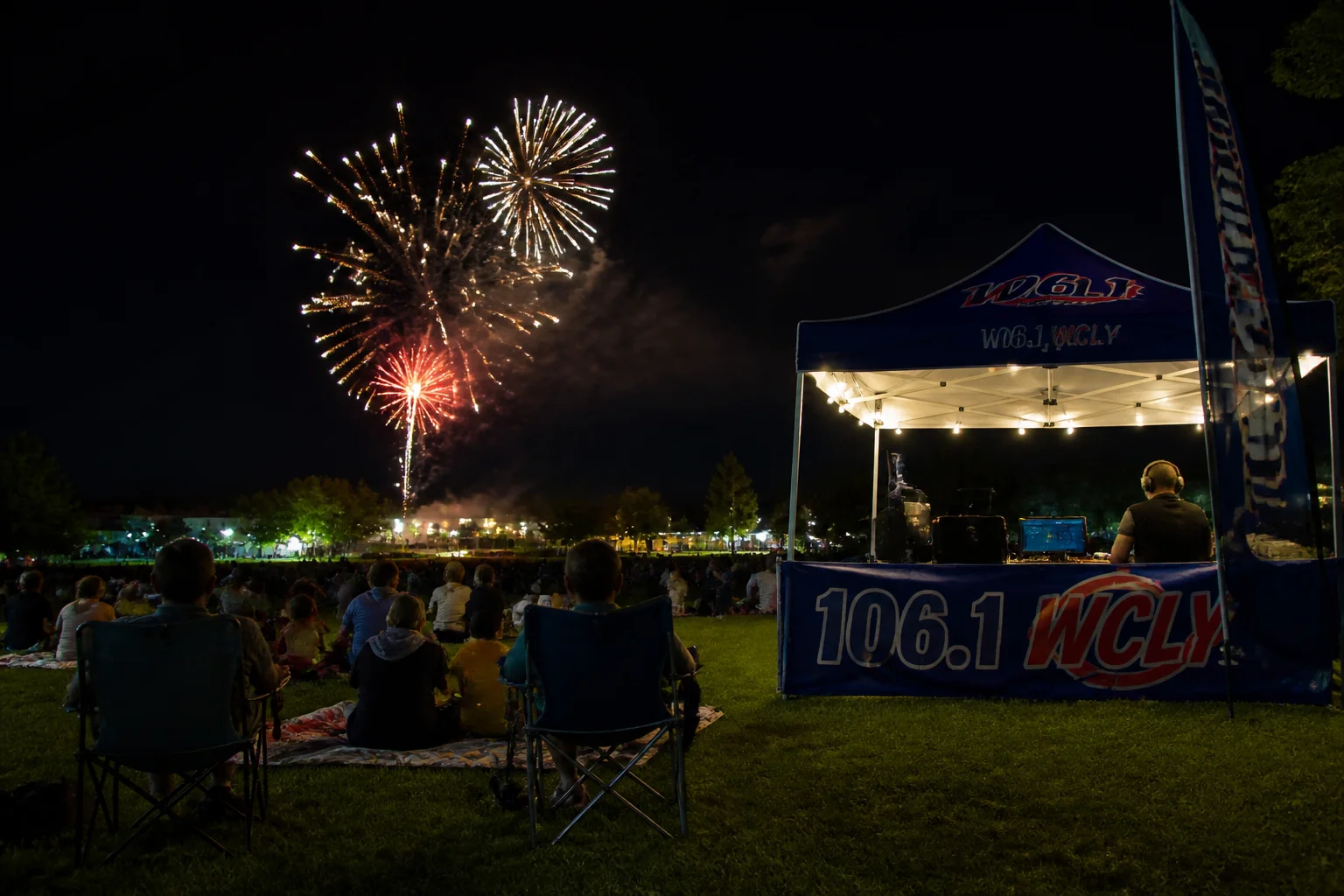 Community Fourth of July fireworks display over a small-town park at night, families watching with a radio station remote tent lit up nearby